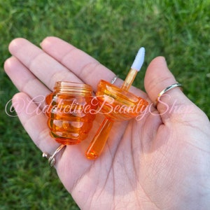 May include: An open, orange-colored lip gloss container with a honeycomb design. The container is made of clear plastic and has a matching applicator wand. The container is held in a hand against a green background.