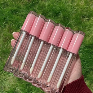 Puede incluir: Seis tubos transparentes de brillo de labios con tapas rosas y aplicadores blancos. Los tubos est&aacute;n dispuestos en fila y se sostienen en una mano.