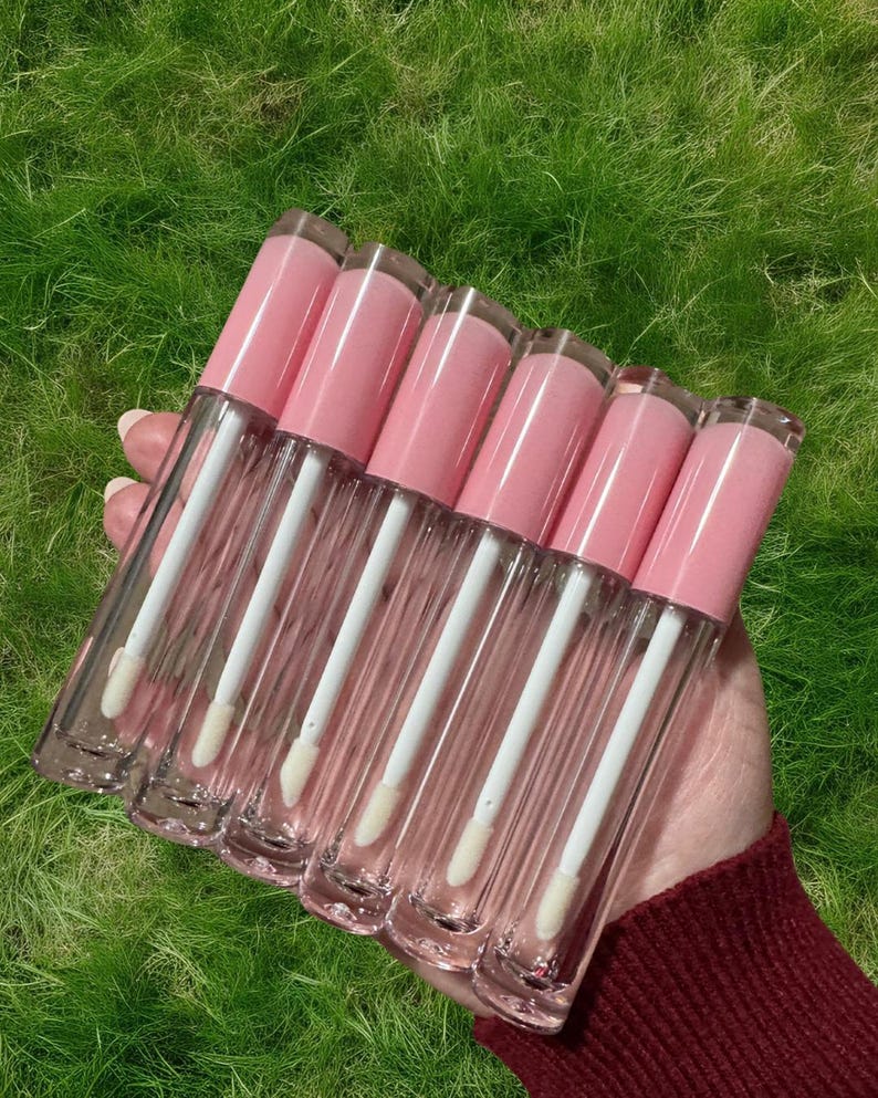 Puede incluir: Seis tubos transparentes de brillo de labios con tapas rosas y aplicadores blancos. Los tubos est&aacute;n dispuestos en fila y se sostienen en una mano.