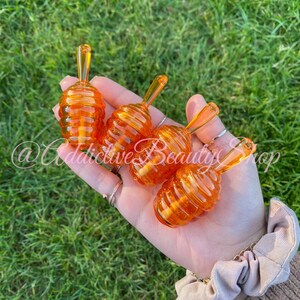 May include: A collection of five orange-tinted, honey dippers, each with a beehive-shaped base and a long, slender handle. The dippers are held in a hand against a backdrop of green grass. The text "@Addictive Beauty Shop" is visible.