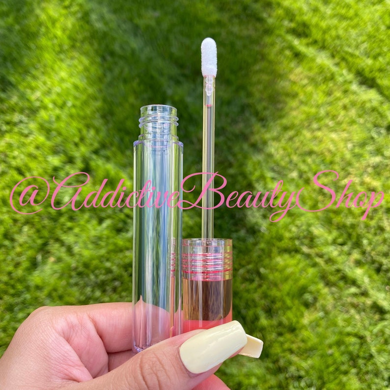 K&ouml;nnte beinhalten: Ein transparentes Lipgloss-R&ouml;hrchen mit einem rosa und silbernen Deckel und einem wei&szlig;en Applikator in Form eines Hirschfu&szlig;es. Das R&ouml;hrchen ist teilweise mit einer klaren Fl&uuml;ssigkeit gef&uuml;llt.
