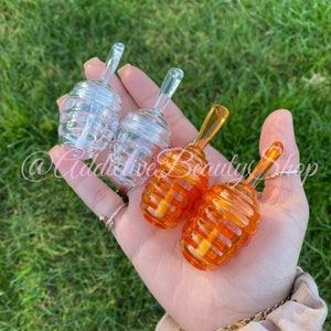 Può includere: Dosatori di miele in vetro trasparente e arancione con un design a nido d'ape. I dosatori sono tenuti in mano su uno sfondo verde. Il testo "@Addictive Beauty Shop" è visibile nell'immagine.