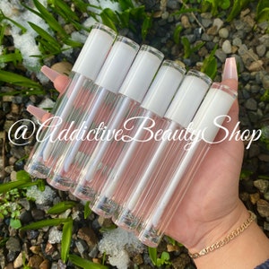 Puede incluir: Un conjunto de siete tubos de brillo de labios transparentes con tapas blancas. Los tubos est&aacute;n dispuestos en forma de abanico y se sostienen en una mano. El texto "@Addictive Beauty Shop" es visible en la imagen.