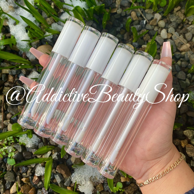 Puede incluir: Un conjunto de siete tubos de brillo de labios transparentes con tapas blancas. Los tubos est&aacute;n dispuestos en forma de abanico y se sostienen en una mano. El texto "@Addictive Beauty Shop" es visible en la imagen.
