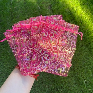 May include: A set of pink organza gift bags with gold and purple swirl designs. The bags are made of sheer fabric and have a drawstring closure.
