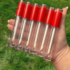 Puede incluir: Seis tubos transparentes de brillo de labios con tapas rojas y aplicadores blancos. Los tubos est&aacute;n dispuestos en una fila, con las tapas mirando al espectador.