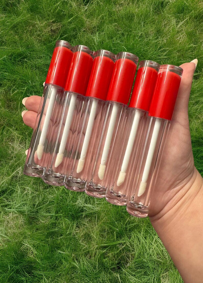 Puede incluir: Seis tubos transparentes de brillo de labios con tapas rojas y aplicadores blancos. Los tubos est&aacute;n dispuestos en una fila, con las tapas mirando al espectador.