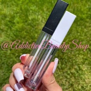 Może przedstawiać: Dwa przezroczyste tuby z błyszczykiem do ust z czarnymi i białymi nakrętkami. Tuby są puste i mają przezroczysty aplikator w postaci wkładki.