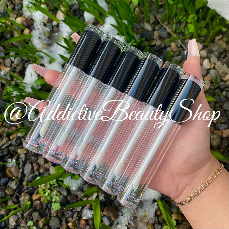 Puede incluir: Un conjunto de seis tubos de brillo de labios transparentes con tapas negras. Los tubos est&aacute;n llenos de un l&iacute;quido transparente ligeramente rosado. Los tubos se sostienen en una mano.