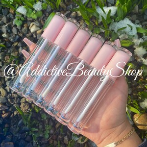 Puede incluir: Un conjunto de seis tubos de brillo labial transparentes con tapas rosadas. Los tubos est&aacute;n dispuestos en forma de abanico y se sostienen en una mano. El texto '@Addictive Beauty Shop' es visible en la imagen.