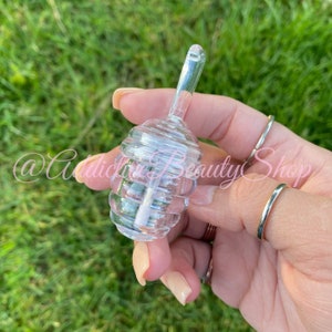 May include: Clear, glass lip gloss container with a spiral design. The container has a long, clear applicator wand. The text "@AddictiveBeautyShop" is visible in the image.