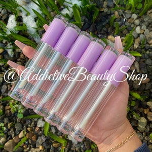 Puede incluir: Un conjunto de ocho tubos de brillo labial transparentes con tapas de color lavanda. Los tubos est&aacute;n dispuestos en forma de abanico y se sostienen en una mano. El texto "@Addictive Beauty Shop" es visible en la imagen.
