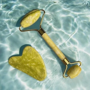 May include: A jade facial roller and a heart-shaped gua sha tool. The roller has a gold-colored metal frame and a handle with a light green, marbled stone. The gua sha tool is a similar light green color. Both are on a blue, rippled surface.