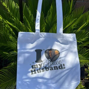 Op de afbeelding: Witte draagtas met de tekst "I my Space Husband!" in zwart en een hartvormige afbeelding van een man. De tas heeft twee witte handvatten en is gemaakt van een lichtgekleurde stof.