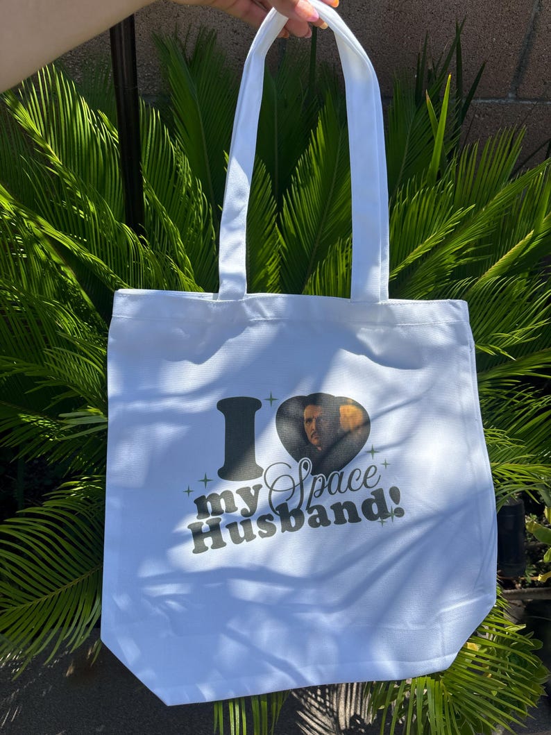 Op de afbeelding: Witte draagtas met de tekst "I my Space Husband!" in zwart en een hartvormige afbeelding van een man. De tas heeft twee witte handvatten en is gemaakt van een lichtgekleurde stof.