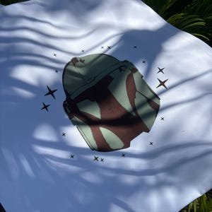 Op de afbeelding: Witte draagtas met een groen en bruin helmdesign en steraccenten. De tas is gemaakt van een lichtgekleurde stof en heeft een eenvoudig handvatontwerp. De afbeelding is buiten gemaakt met natuurlijk licht.