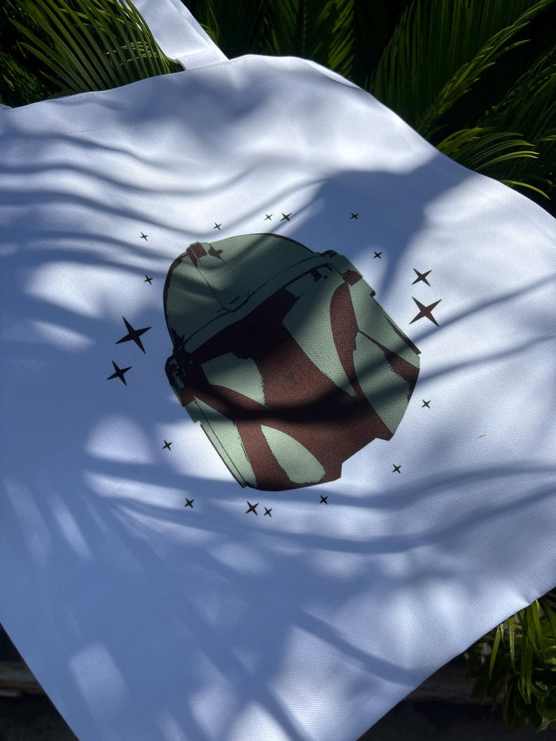 Op de afbeelding: Witte draagtas met een groen en bruin helmdesign en steraccenten. De tas is gemaakt van een lichtgekleurde stof en heeft een eenvoudig handvatontwerp. De afbeelding is buiten gemaakt met natuurlijk licht.