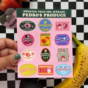 Peut inclure: Une planche d'autocollants colorés avec le texte "Pedro's Produce" et des motifs sur le thème des fruits. Les autocollants sont sur un fond rose, avec un motif de damier en arrière-plan. Une main tient la planche d'autocollants, avec une banane, un avocat, une fraise et une orange dans le cadre.