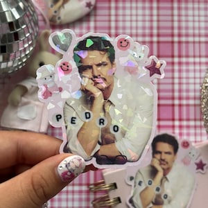 May include: A holographic sticker with a portrait of a man, the name "PEDRO" spelled out in letter beads. The sticker is decorated with small holographic hearts, stars, and smiley faces. Other stickers and a disco ball are visible in the background.
