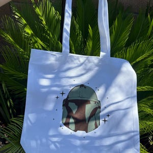 Op de afbeelding: Witte draagtas met een groen en bruin helmdesign en steraccenten. De tas heeft twee witte handvatten en is gemaakt van een stevige stof, ideaal voor het dragen van boodschappen of persoonlijke spullen.