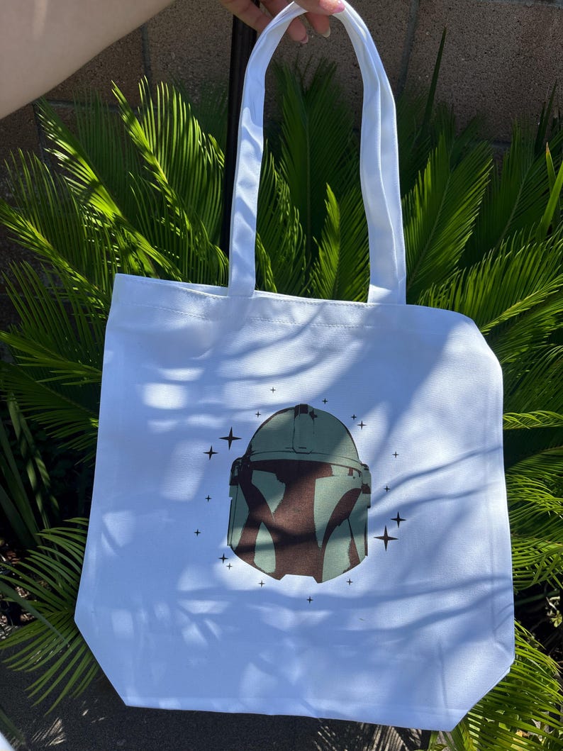 Op de afbeelding: Witte draagtas met een groen en bruin helmdesign en steraccenten. De tas heeft twee witte handvatten en is gemaakt van een stevige stof, ideaal voor het dragen van boodschappen of persoonlijke spullen.