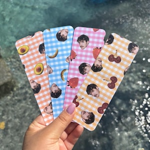 May include: Four colorful bookmarks with a gingham pattern and a portrait. Each bookmark features a different fruit: avocado, banana, strawberry, and cherries. The background is a blurred view of water.