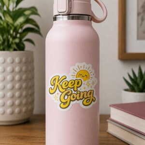 Puede incluir: Una botella de agua de color rosa claro mate con tapa abatible y asa de transporte. Una pegatina en la parte frontal muestra un sol amarillo con cara sonriente y el texto "Keep Going" en fuente retro, rodeado de pequeños iconos de estrellas.