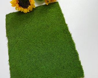 Puede incluir: Un trozo rectangular de musgo artificial verde brillante, con una superficie densa y texturizada. El musgo se coloca sobre un fondo blanco, con unos girasoles amarillos en la esquina superior izquierda. El musgo parece ser un elemento decorativo.