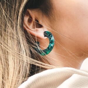 May include: A close-up of a person's ear wearing a pair of green and white hoop earrings. The earrings are made of a marbled material and have a slightly irregular shape.