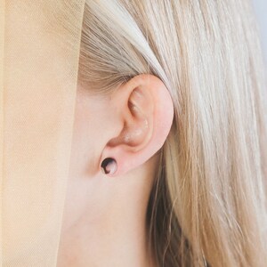 May include: Close-up of an ear with a dark brown and light brown circular earring. The earring is a plug style. The person has blonde hair and is partially obscured by a sheer, beige fabric.