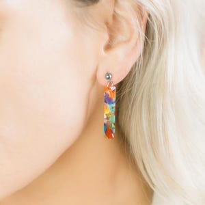 May include: A pair of silver stud earrings with a colourful rectangular dangle. The dangle is made of a translucent material with a variety of colours, including orange, red, blue, green, and yellow.