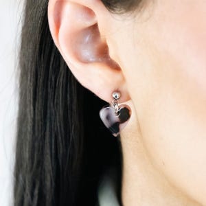 May include: A heart-shaped earring with a silver post. The earring features a translucent, marbled design in grey and brown tones. The earring is suspended from a person's ear, close up.