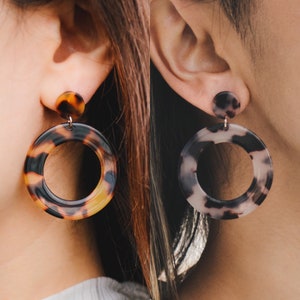 May include: A pair of large, round hoop earrings with a tortoiseshell pattern. The earrings are made of a brown and black resin material. One earring is brown and black, and the other is white and black.