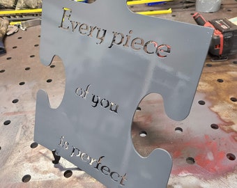 May include: A gray metal puzzle piece with the words "Every piece of you is perfect" cut out. The metal art piece is displayed on a metal table with tools and supplies in the background.