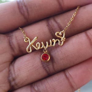 May include: A gold-colored wire name necklace spelling "Kevin" with a small red gemstone pendant. The necklace features a delicate chain and a heart-shaped design element. The name is crafted in a cursive style.