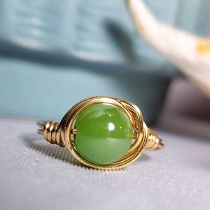 May include: A gold wire wrapped ring with a green gemstone. The ring is on a white surface.