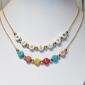 May include: Gold chain necklace with two layers of beads. The top layer has white beads with the name "JOSLYNN" spelled out in gold letters. The bottom layer has colorful beads with the name "JAELYNN" spelled out in gold letters.