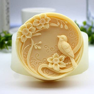 Puede incluir: Un molde de silicona redondo para hacer jabón o velas. El molde presenta un diseño detallado de un pájaro posado en una rama con flores.