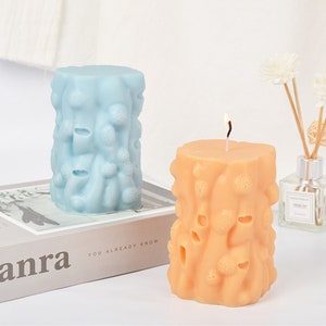 May include: Two decorative pillar candles with a textured surface resembling tree bark. The candle on the left is light blue and the candle on the right is orange.