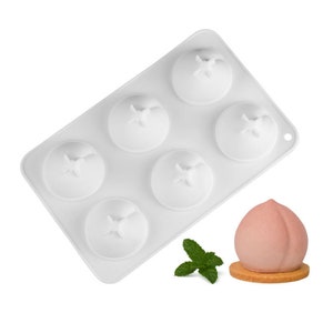 Könnte beinhalten: Weiße Silikonform mit sechs pfirsichförmigen Vertiefungen. Die Form befindet sich auf weißem Hintergrund. Ein rosafarbener pfirsichförmiger Nachtisch sitzt auf einem runden, braunen Keks.