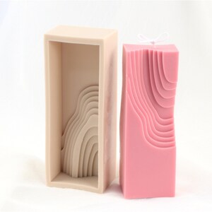 May include: A pink candle with a wavy, layered design in a rectangular silicone mold. The mold is beige and has a similar wavy design.