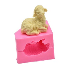 May include: Pink silicone mould for baking a lamb-shaped cake. The mould has a detailed design of a sitting lamb with a fluffy texture.