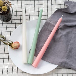 May include: Three taper candles in white, green, and pink rest on a white plate with a dried rose. A gray cloth and a black and white checkered tablecloth are in the background. A small vase with yellow flowers is also visible.