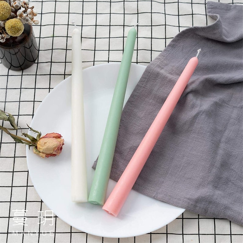May include: Three taper candles in white, green, and pink rest on a white plate with a dried rose. A gray cloth and a black and white checkered tablecloth are in the background. A small vase with yellow flowers is also visible.