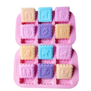 Pode incluir: Forma de silicone rosa com 12 cavidades quadradas, cada uma com um signo do zodíaco diferente. A forma está cheia de sabonetes quadrados coloridos, cada um com um símbolo do zodíaco.