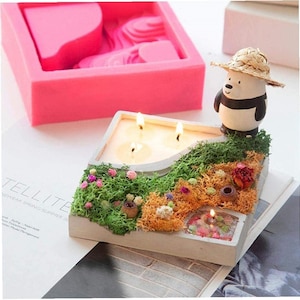 Könnte beinhalten: Ein weißer Beton-Kerzenhalter mit einer Panda-Figur, die einen Strohhut trägt. Der Halter hat einen geschwungenen Weg mit drei brennenden Kerzen und einen kleinen Teich mit einer brennenden Kerze. Der Halter ist von grünem und braunem Moos und kleinen Blumen umgeben.