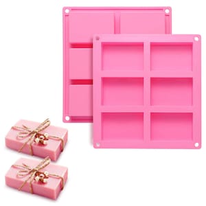 Pode incluir: Dois moldes de silicone rosa para fazer barras de sabão. Cada molde tem seis cavidades retangulares. Duas barras de sabão rosa são envoltas em barbante e decoradas com pequenas flores brancas.