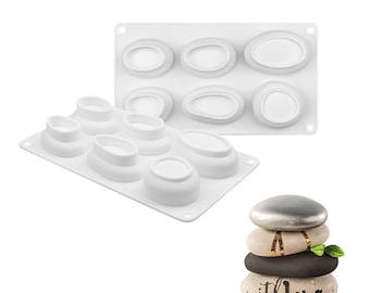 Puede incluir: Moldes de silicona blancos en dos diseños diferentes, uno con formas ovaladas y otro con formas de piedras apiladas. También se muestra una pila de piedras decorativas con la inscripción "with love".
