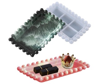 May include: Two rectangular trays with a beaded border. One tray is black with a green marble pattern, the other is white. A pink tray with a beaded border is filled with two black lipstick tubes and a gold crown-shaped trinket holder.