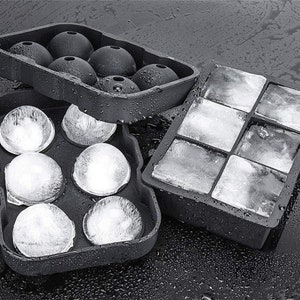 Puede incluir: Dos bandejas de cubitos de hielo de silicona negra. Una bandeja tiene seis moldes de hielo redondos y la otra tiene nueve moldes de hielo cuadrados. Ambas bandejas están llenas de hielo.